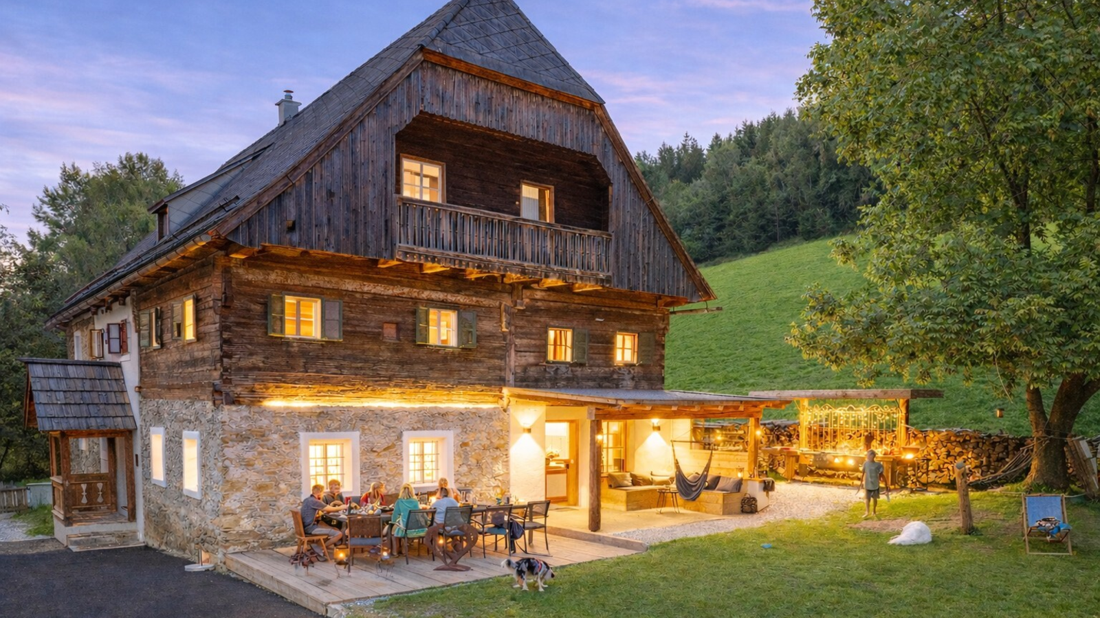 Exterior of the farmhouse featuring an illuminated terrace, outdoor dining area, and a covered lounge in the garden.