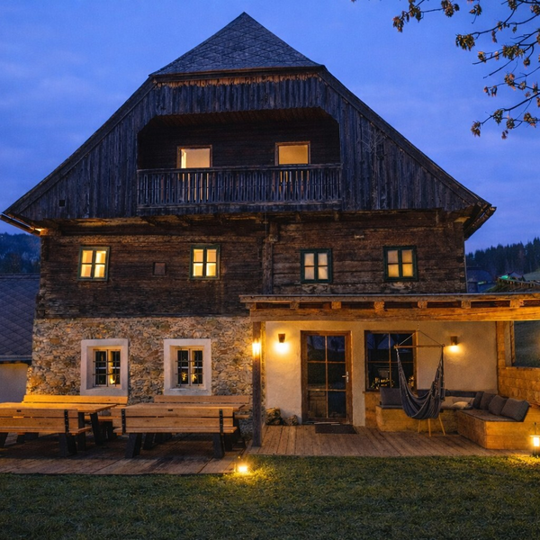 Exterior view of the farmhouse at dusk, featuring illuminated windows, a terrace with benches, and a covered porch with a hammock.