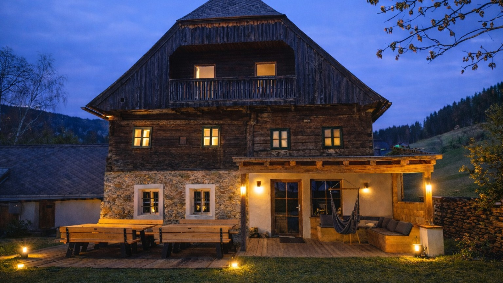 Exterior view of the farmhouse at dusk, featuring illuminated windows, a terrace with benches, and a covered porch with a hammock.