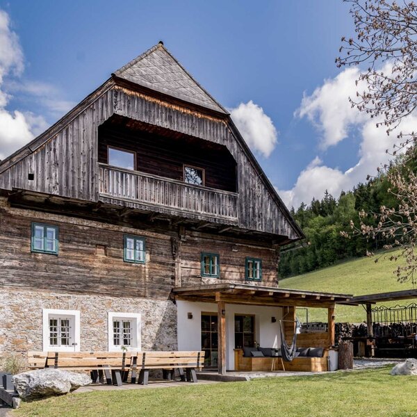 The exterior of the farmhouse featuring traditional wood and stone facade, a balcony, and a covered terrace with garden seating.