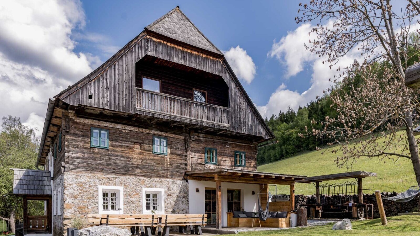 The exterior of the farmhouse featuring traditional wood and stone facade, a balcony, and a covered terrace with garden seating.