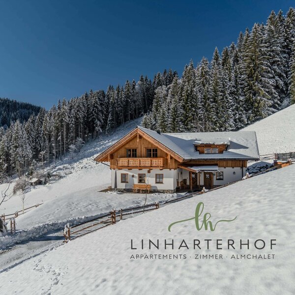 Exterior view of the farmhouse, featuring a traditional wooden balcony, surrounded by snow-covered mountains and pine forests under a clear sky.