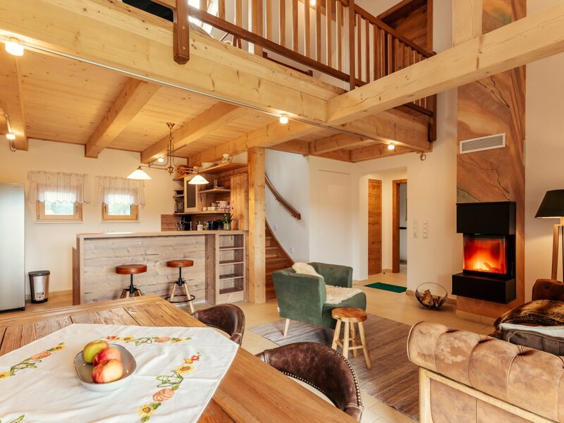 The open-plan living area of the Alpin Chalet, featuring a kitchen with a bar, a dining table, a fireplace, and seating under a wooden ceiling.