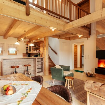 The open-plan living area of the Alpin Chalet, featuring a kitchen with a bar, a dining table, a fireplace, and seating under a wooden ceiling.