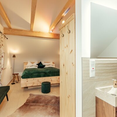 Bedroom with a double bed, wooden beams, and an adjacent bathroom area featuring a sink and backlit mirror.