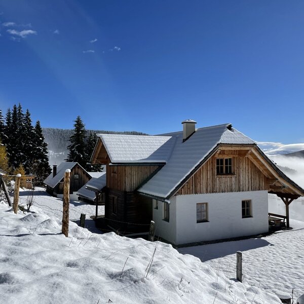 The farmhouse features a snow-covered roof and wooden gables, set amidst snow-dusted pine trees with a distant view over a misty valley.