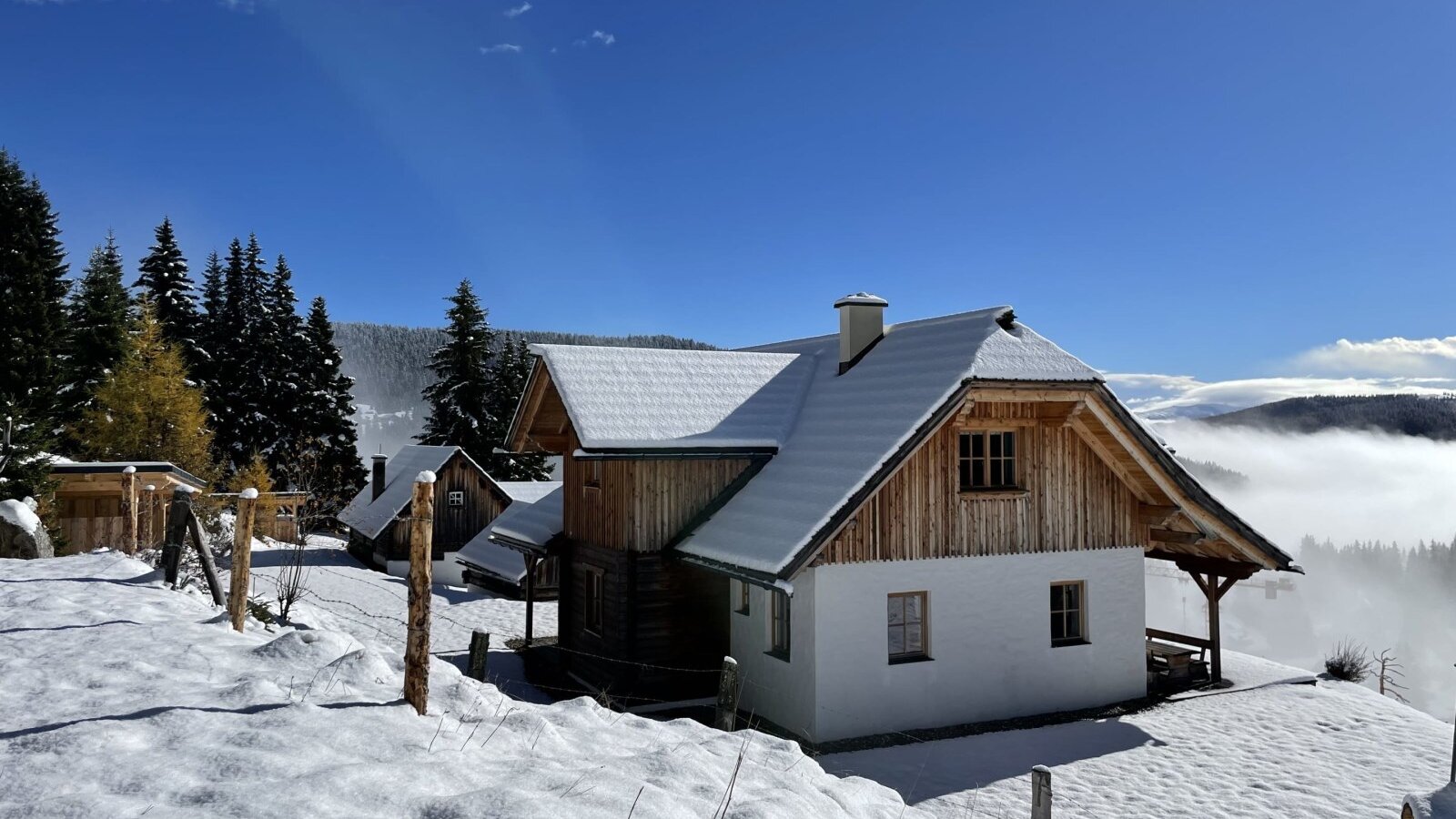 The farmhouse features a snow-covered roof and wooden gables, set amidst snow-dusted pine trees with a distant view over a misty valley.