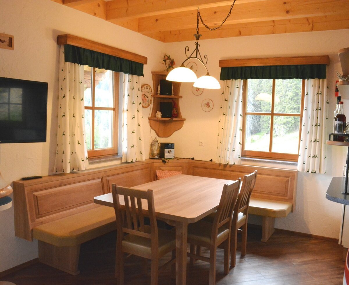 The farmhouse's dining area features a wooden table with built-in bench seating and chairs, a wall-mounted TV, and windows overlooking the surrounding greenery.