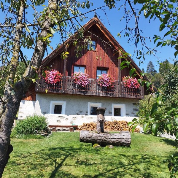 The inviting farmhouse with a flower-adorned balcony and a green lawn featuring a rustic wooden fountain.