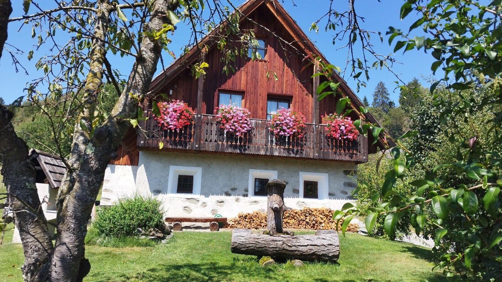 The inviting farmhouse with a flower-adorned balcony and a green lawn featuring a rustic wooden fountain.