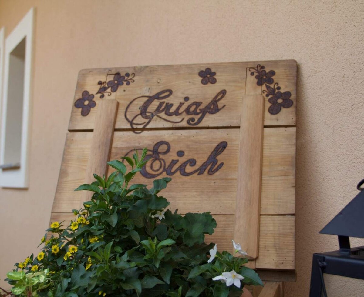 Wooden welcome sign 'Griaß Eich' with flowers at the farmhouse entrance.