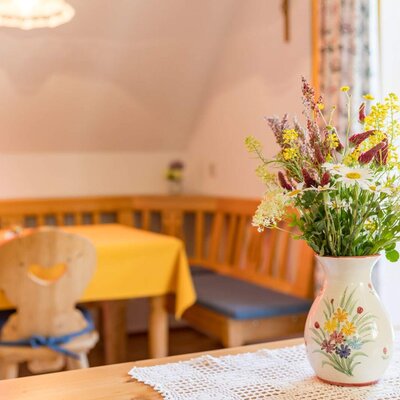 The seating area in the eat-in kitchen of the apartment on the farmhouse, with a wooden table, corner bench, and a flower arrangement.