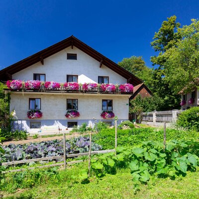 The farm house exterior features a balcony with vibrant flower boxes and a garden with various plants in the foreground.