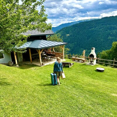 The farmhouse features a covered outdoor dining area, a grassy lawn, an outdoor grill, and mountain views as a guest arrives.