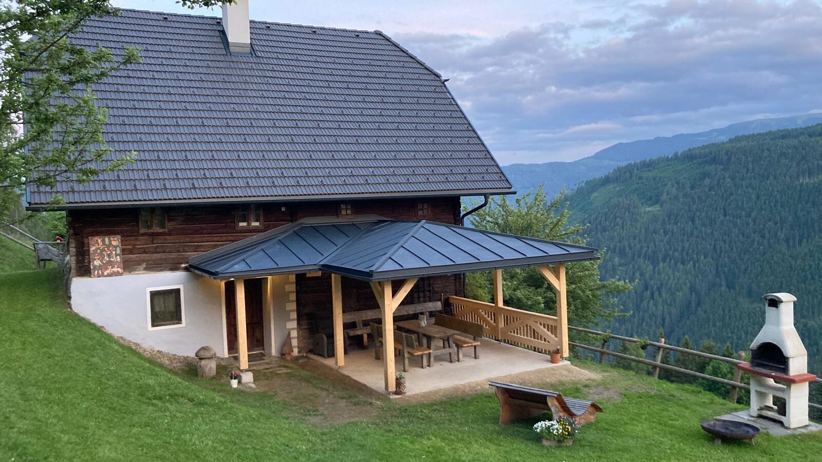 The farmhouse exterior includes a covered patio with wooden seating, an outdoor barbecue grill, and views of the surrounding mountains and forests.