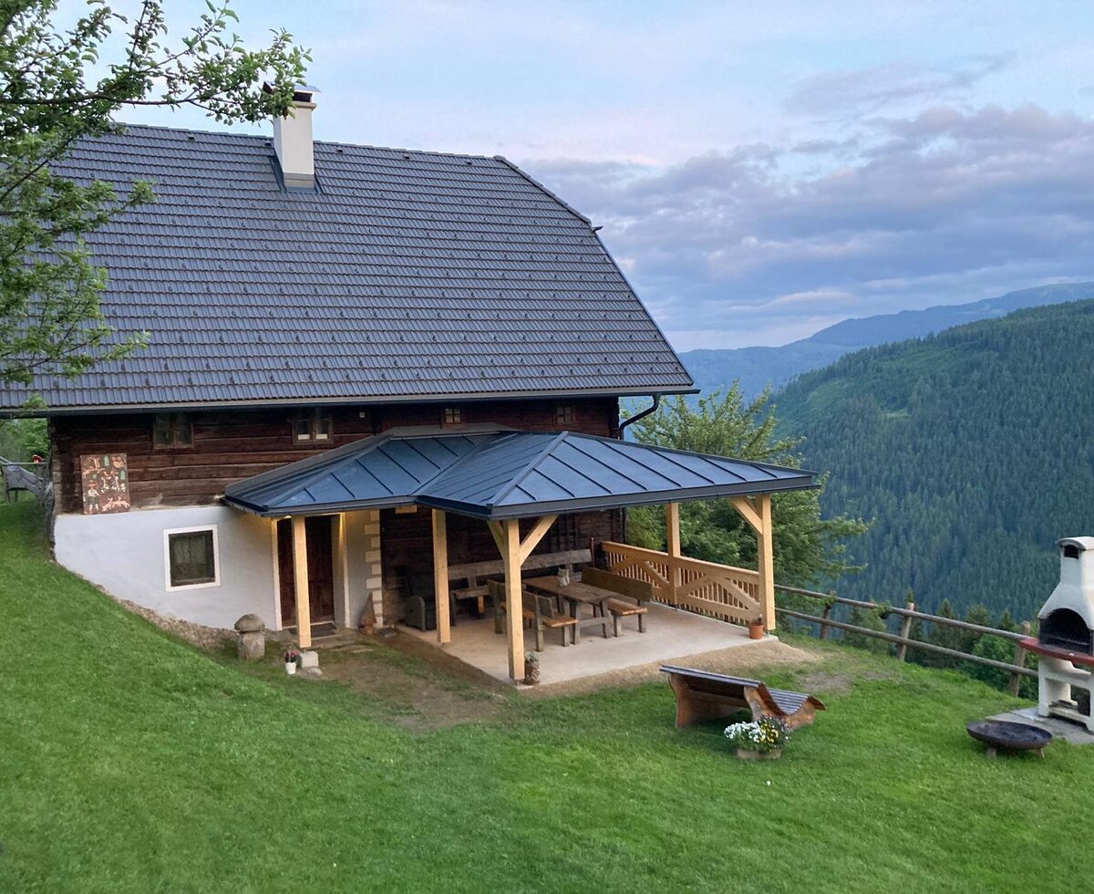 The farmhouse exterior includes a covered patio with wooden seating, an outdoor barbecue grill, and views of the surrounding mountains and forests.