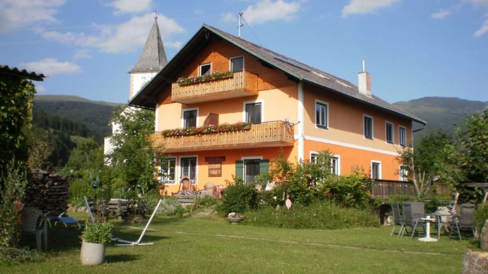 The two-story farm house features balconies with flower boxes, a front garden with outdoor seating, and views of the surrounding mountains and a church steeple.