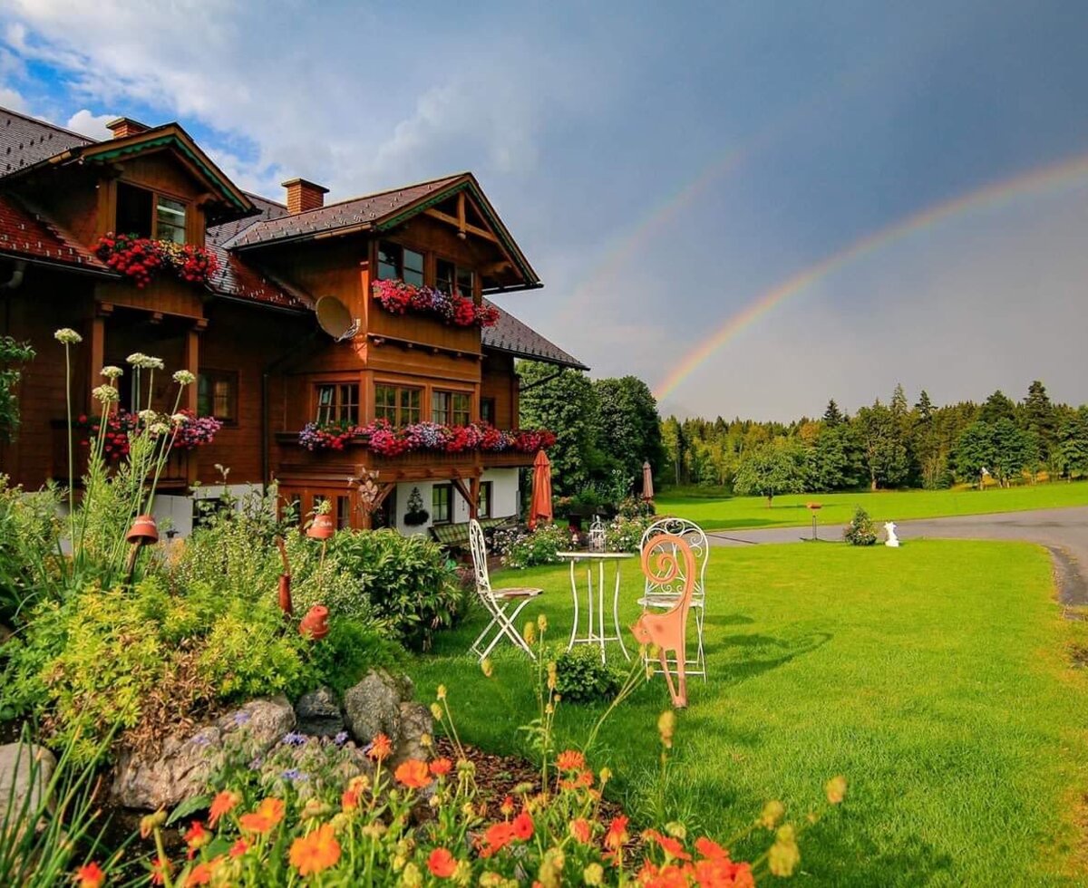 The farmhouse with its blooming garden and lawn, surrounded by meadows and forest, with a rainbow in the sky.