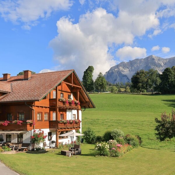 The inviting farmhouse featuring flower-decked balconies, surrounded by lush green meadows and a scenic mountain backdrop.