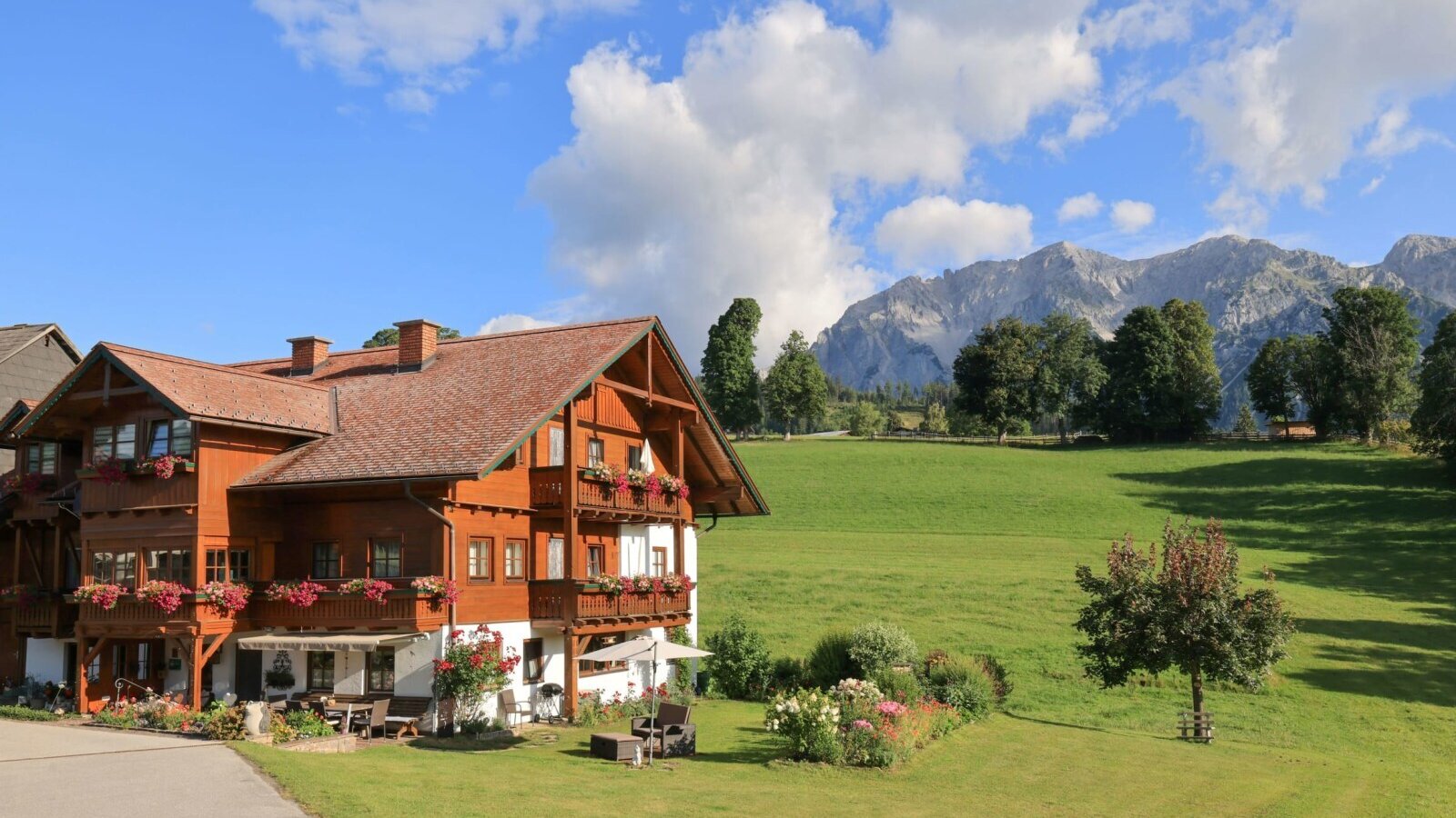 The inviting farmhouse featuring flower-decked balconies, surrounded by lush green meadows and a scenic mountain backdrop.