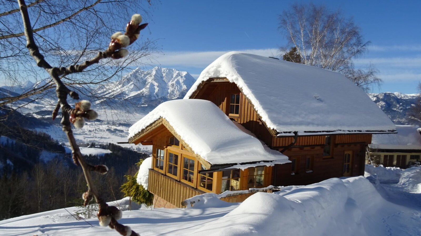 The snow-covered farmhouse with wooden walls and mountain views under a blue sky.