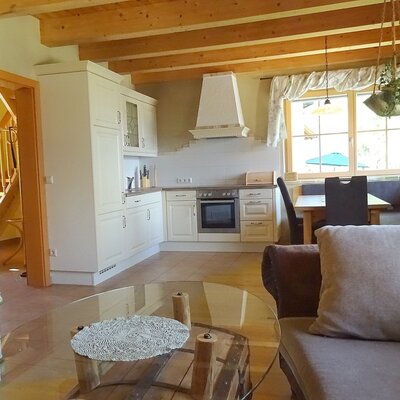 The living area of the farmhouse with an open kitchen, dining area, sofa, and tiled stove under wooden beams.