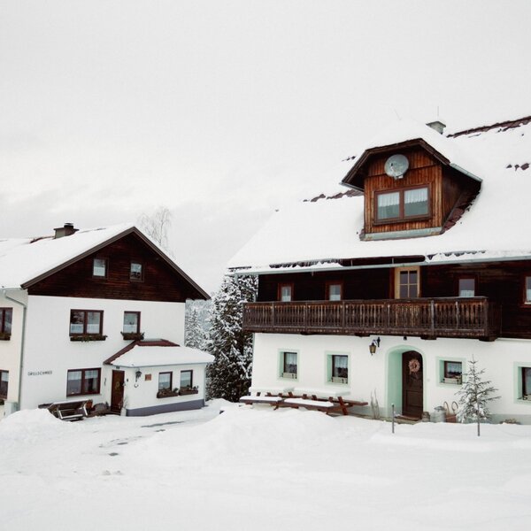 The snow-covered exterior of the farm house and an adjacent building in a winter setting, featuring wooden balconies and dormer windows. The snow-covered exterior of the farm house and an adjacent building in a winter setting, featuring wooden balconies and dormer windows.