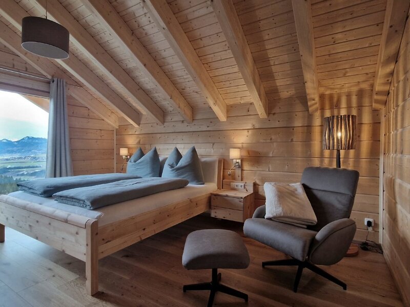 Double bedroom in the farmhouse with a wooden bed, armchair, and mountain view through the large window.