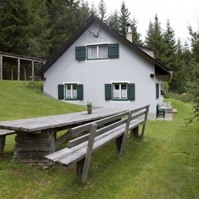 The outdoor area of the alpine hut with a wooden table and benches, surrounded by forest. cMiriamRaneburger
