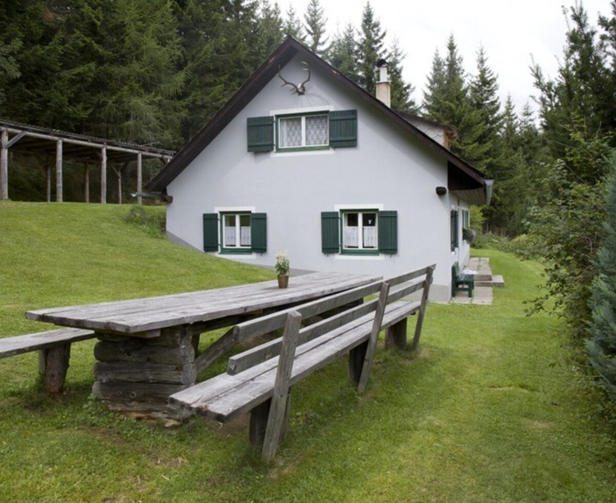 The outdoor area of the alpine hut with a wooden table and benches, surrounded by forest. cMiriamRaneburger