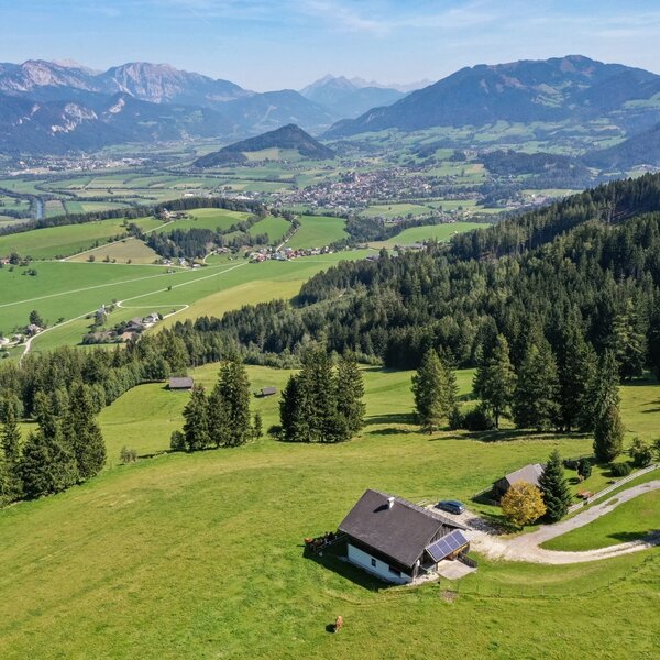 The farmhouse, featuring solar panels, is situated on a green hillside with surrounding trees, offering panoramic views of the valley, a town, and mountains.