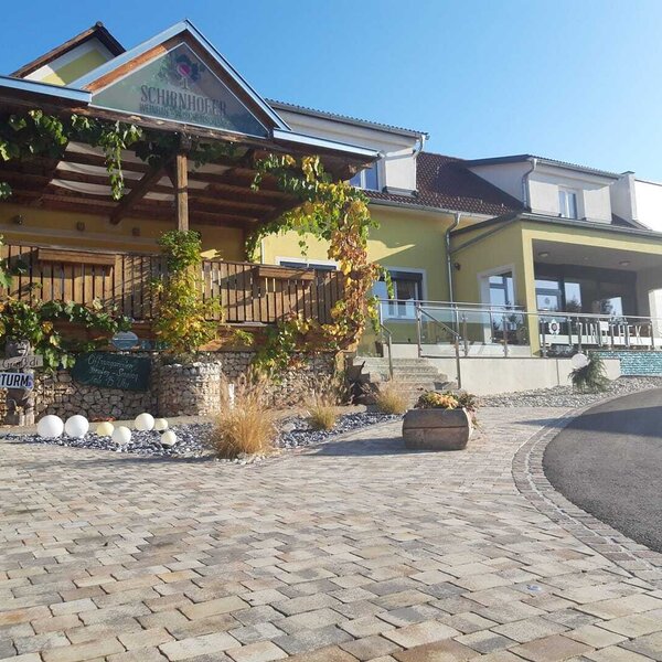 The Schirnhofer farmhouse exterior, featuring a covered wooden terrace with climbing vines, a paved entrance, and a separate sheltered outdoor seating area.