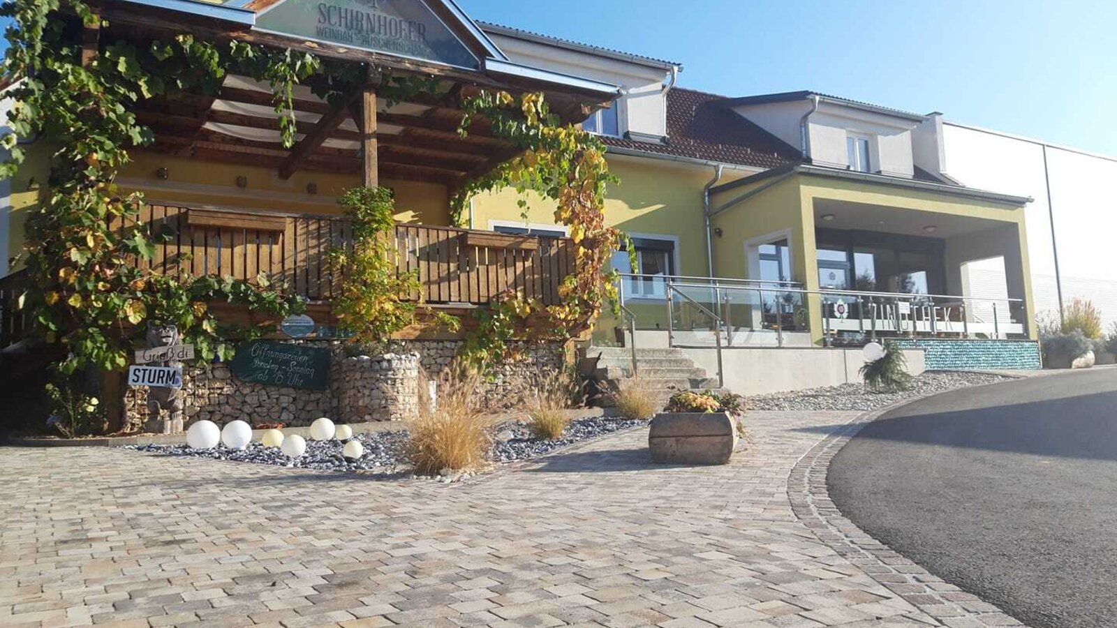 The Schirnhofer farmhouse exterior, featuring a covered wooden terrace with climbing vines, a paved entrance, and a separate sheltered outdoor seating area.