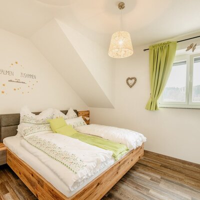 Double bedroom in the farmhouse featuring a wooden bed frame, a bedside table with a lamp, a window with green curtains, and a wooden floor.