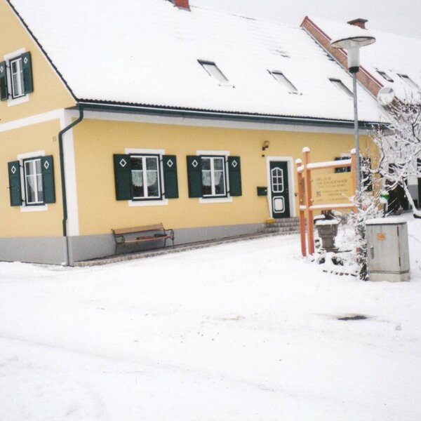 The yellow farmhouse with green window shutters and a snow-covered roof, providing an exterior view of the property in winter.