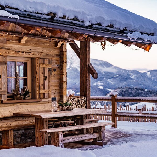The farmhouse's snow-covered outdoor terrace features a wooden table and benches, decorative lighting, and a view of the surrounding mountains.