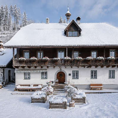 The traditional farmhouse is beautifully covered in snow, featuring a charming wooden balcony and a picturesque, snow-covered front yard.