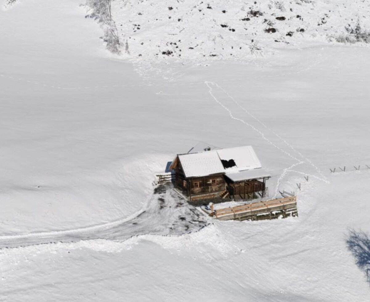 The wooden farmhouse, featuring a snow-covered roof, is situated in a winter landscape with an access path leading to the entrance.