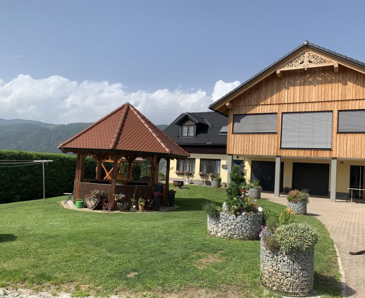 The exterior of the farm house features a garden with a wooden gazebo and gabion planters, alongside a paved driveway and mountain views.