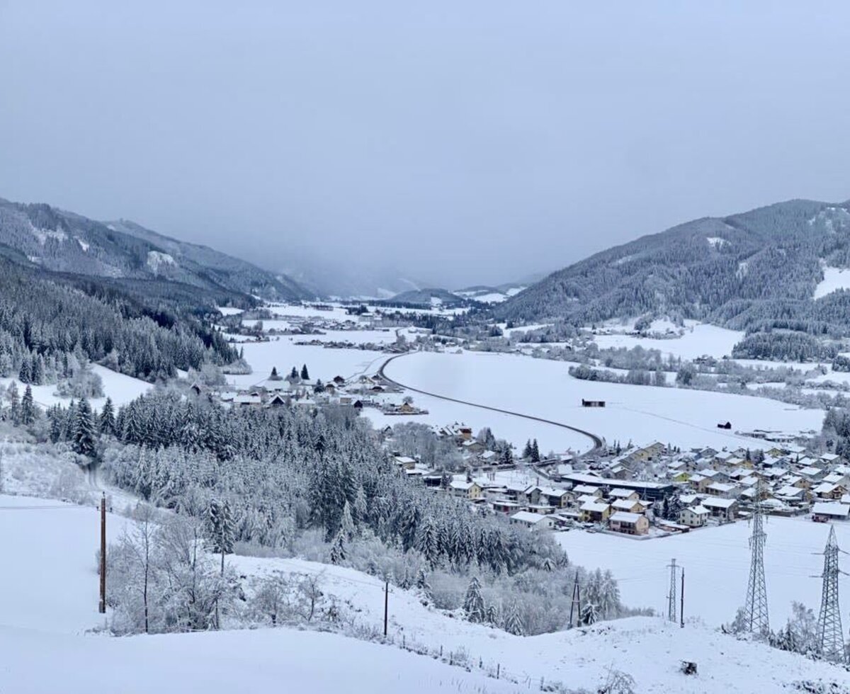View from the farmhouse of the snowy Pölstal valley with mountains, forests, and a village.