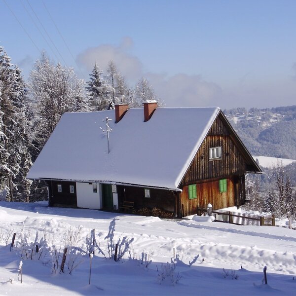 The farm house featuring a wooden exterior and a snow-covered roof, situated in a snowy mountain landscape.