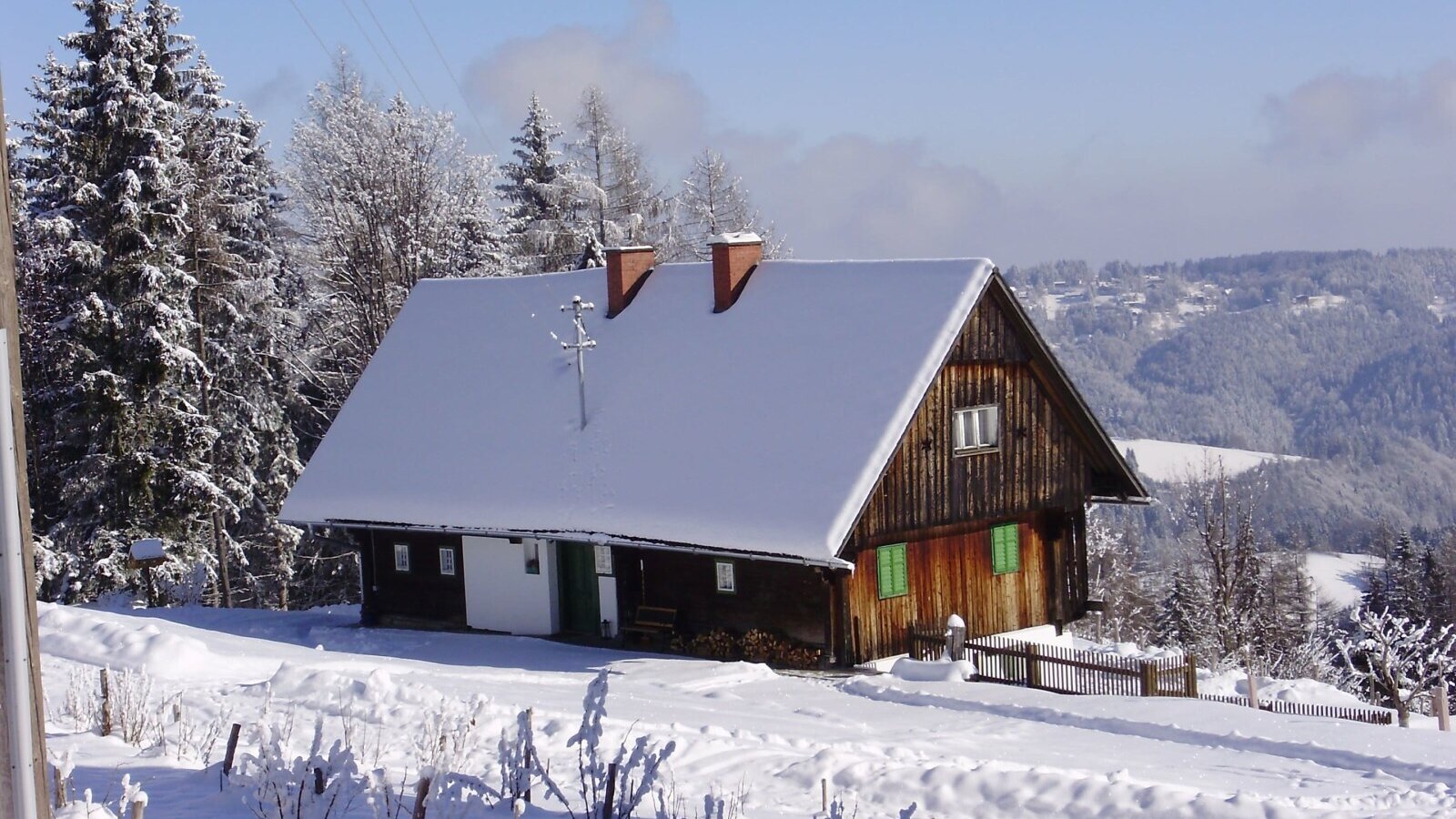 The farm house featuring a wooden exterior and a snow-covered roof, situated in a snowy mountain landscape.