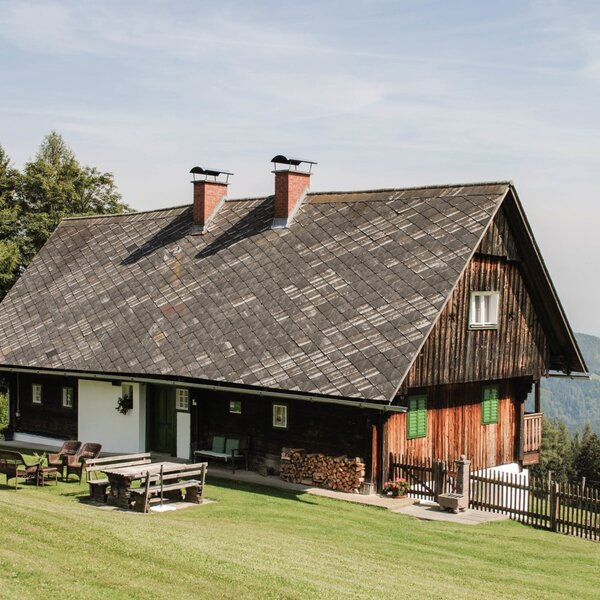 The farmhouse, featuring a wooden facade, a lawn with outdoor seating, a children's swing, and mountain views.