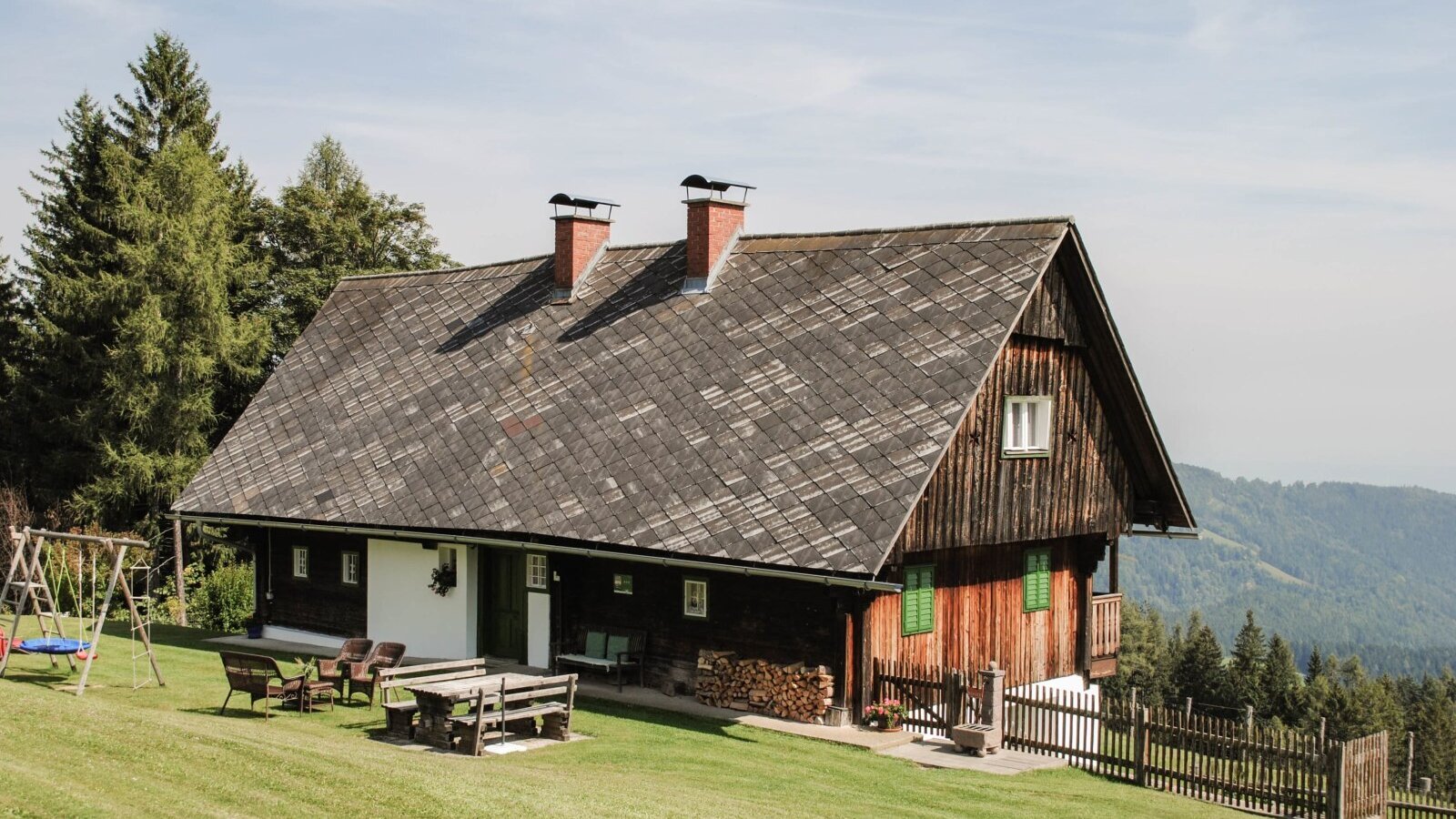 The farmhouse, featuring a wooden facade, a lawn with outdoor seating, a children's swing, and mountain views.