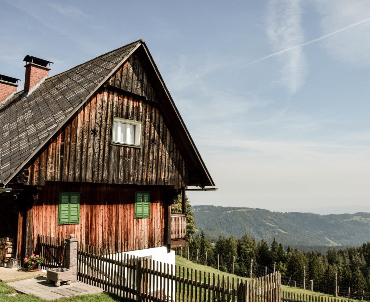 Exterior view of the farmhouse with a wooden facade, traditional roof, and a view of the forested mountain landscape.