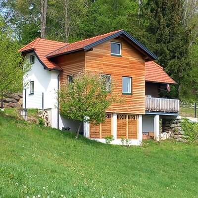 The Farm House features a white and wooden facade, a red tiled roof, and a balcony, situated on a green, grassy slope.