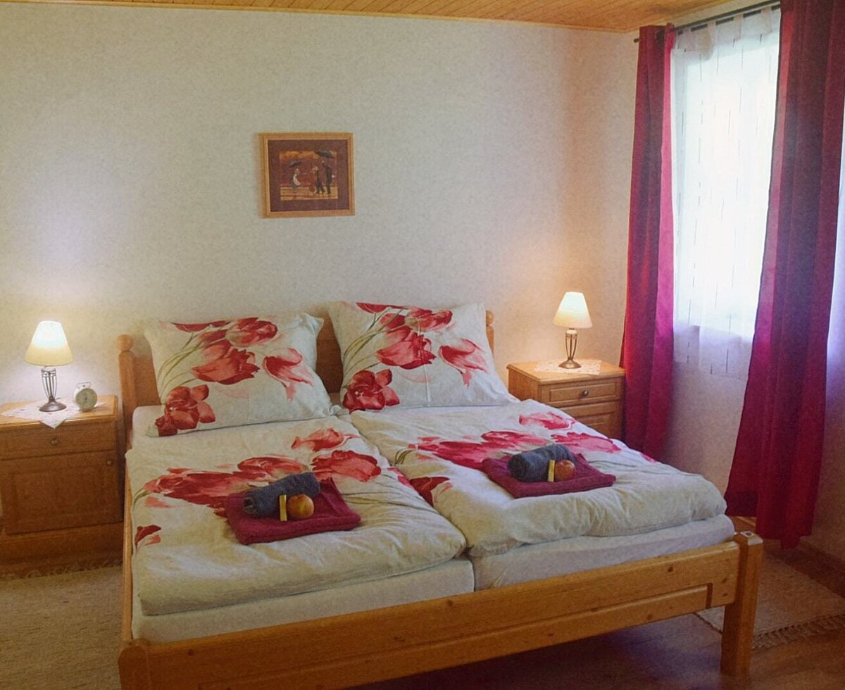 Bedroom in the Farm House featuring two single beds with floral bedding, individual bedside tables with lamps, and a window with red curtains.