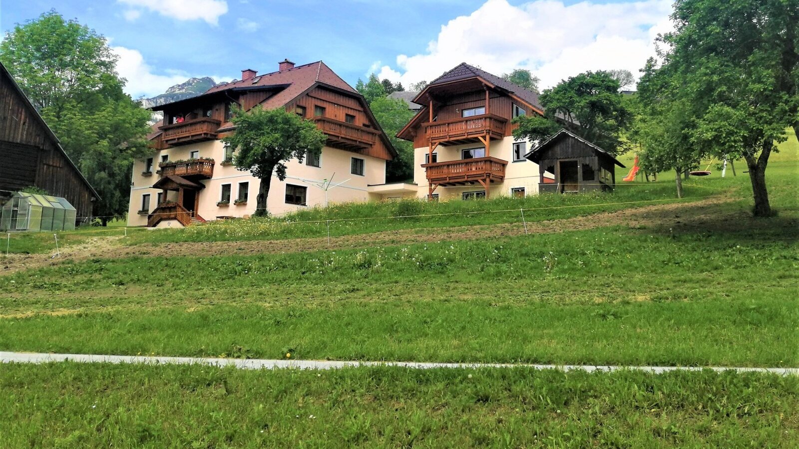 The farmhouse property features two main buildings with multiple balconies, a spacious lawn with a children's play area, and distant views of the Alps.