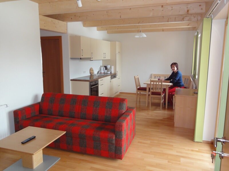 The apartment's open-plan living space combines a sitting area with a sofa, a kitchenette, and a dining area with a table and bench, all under exposed wooden beams.