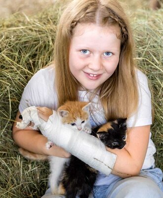 A cheerful young girl cuddles with two adorable little kittens in the hay on the farmhouse.